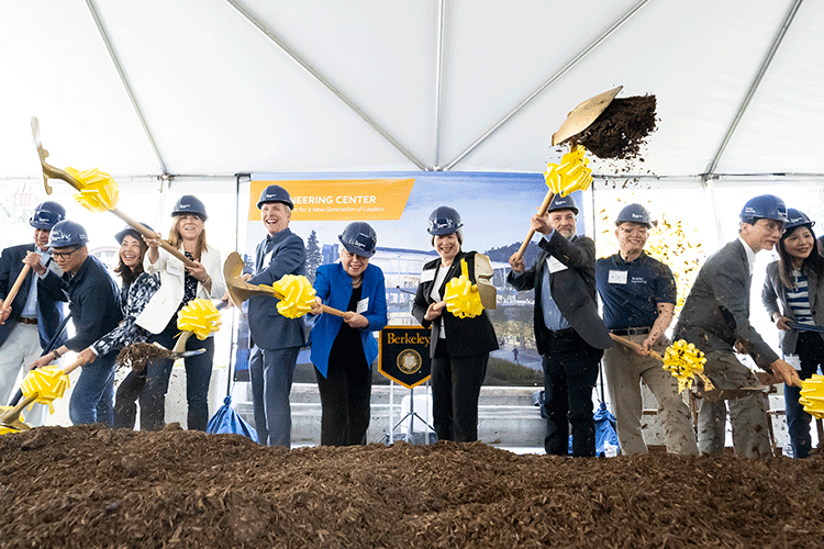 A row of people all wearing hard hats dig shovels into the dirt.