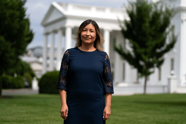 Julie Chavez Rodriguez, wearing a navy blue dress, stands in front of the White House on a sunny summer day