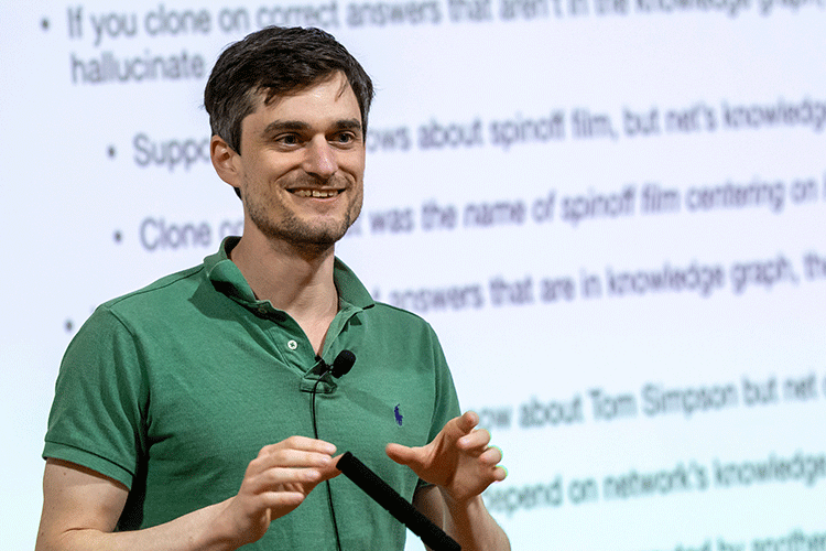 John Schulman stands at a podium in front of a projection of black text on a white background.