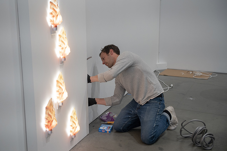 A master of fine arts student kneels on the floor of the Berkeley Art Museum while installing flame-shaped lights on a gallery wall.