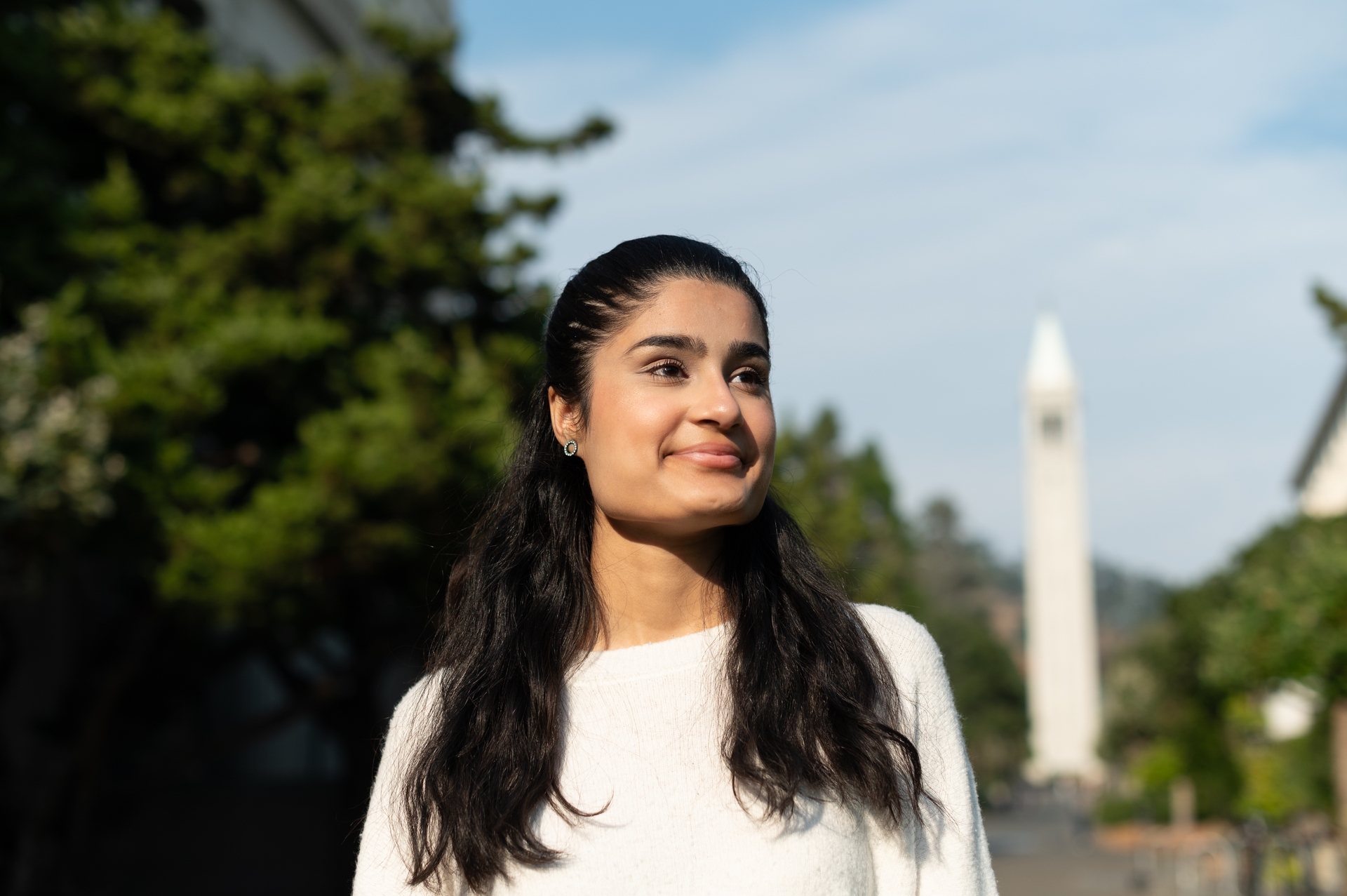 A headshot of Alishba wearing a white blouse, standing outside with the campanile in the background looking towards the distance.