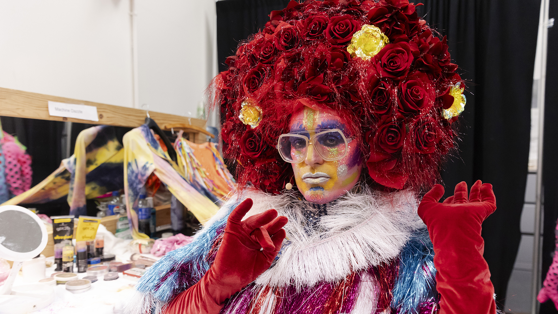 Machine Dazzle, dressed in a huge wig made of bright red and a few yellow fake roses, a red, white and blue tinsel dress and long red velvety gloves with bright makeup covering his face, poses for a portrait