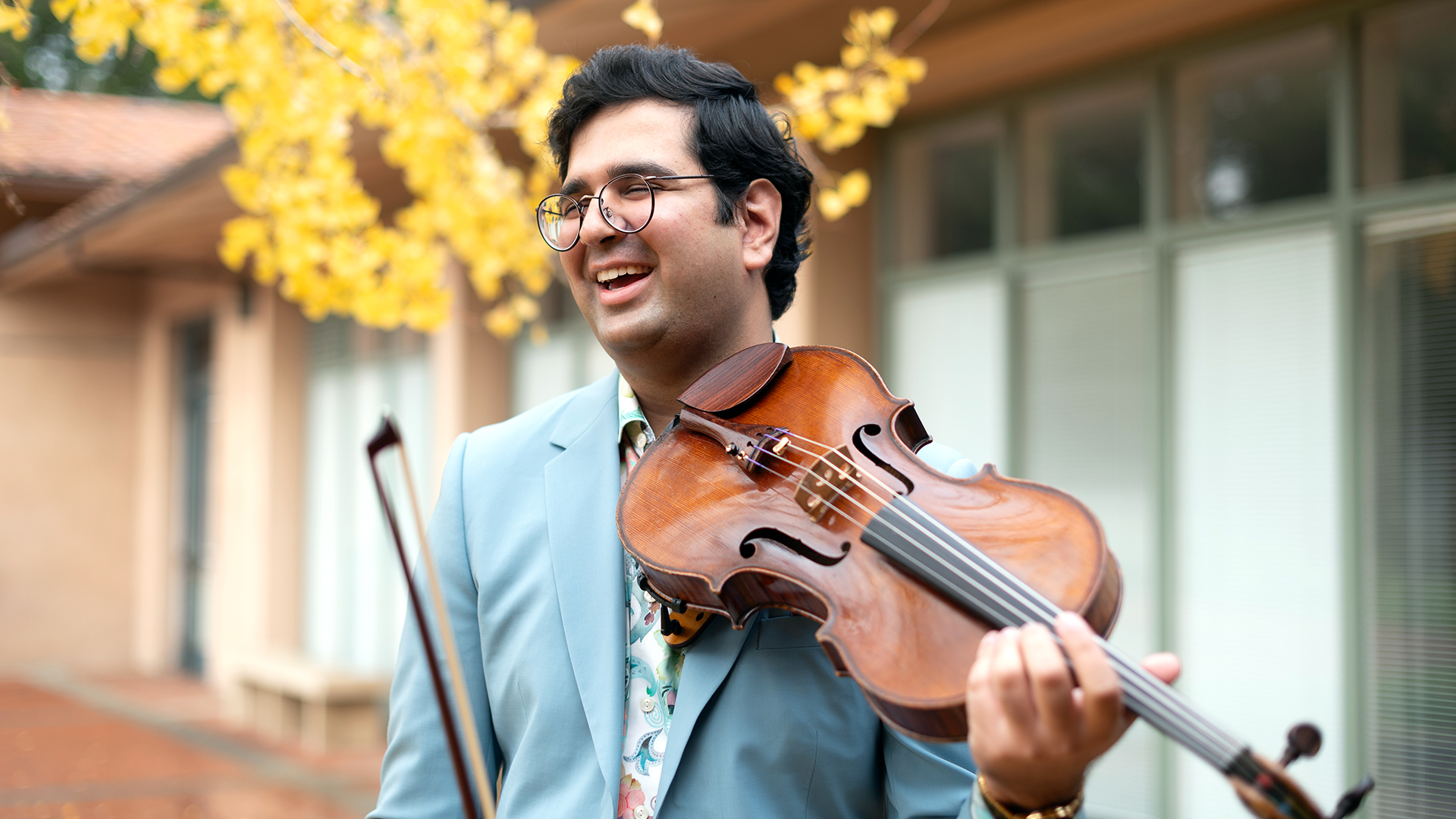 Saagar, with short dark hair and glasses, wearing a powder blue suit, holds a viola to his chin while standing in a courtyard