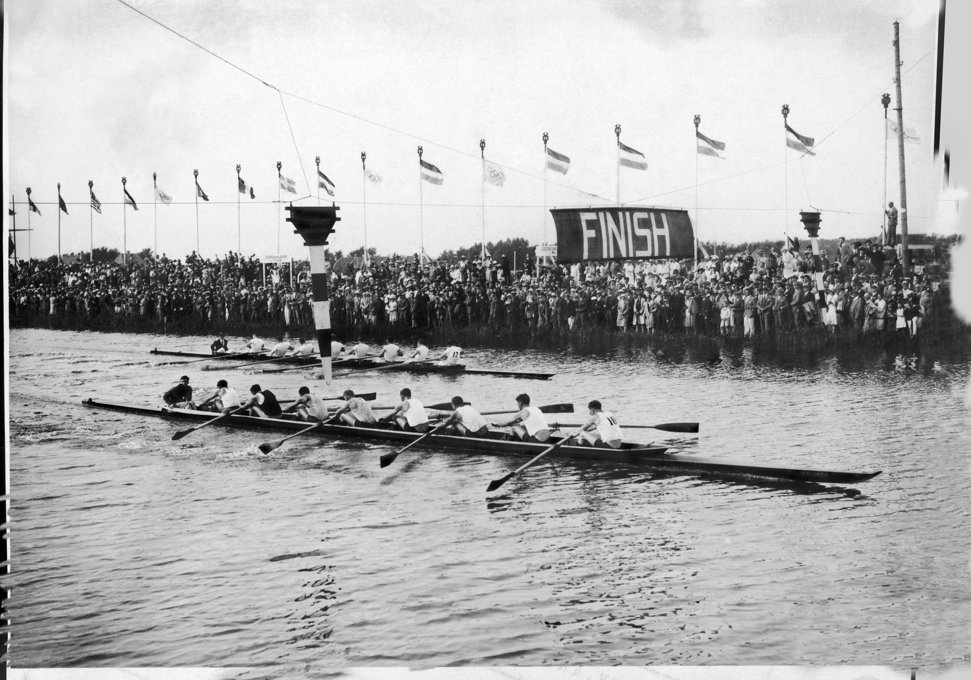 A black and white image of boats being rowed in a race as they cross under a sign that says finish