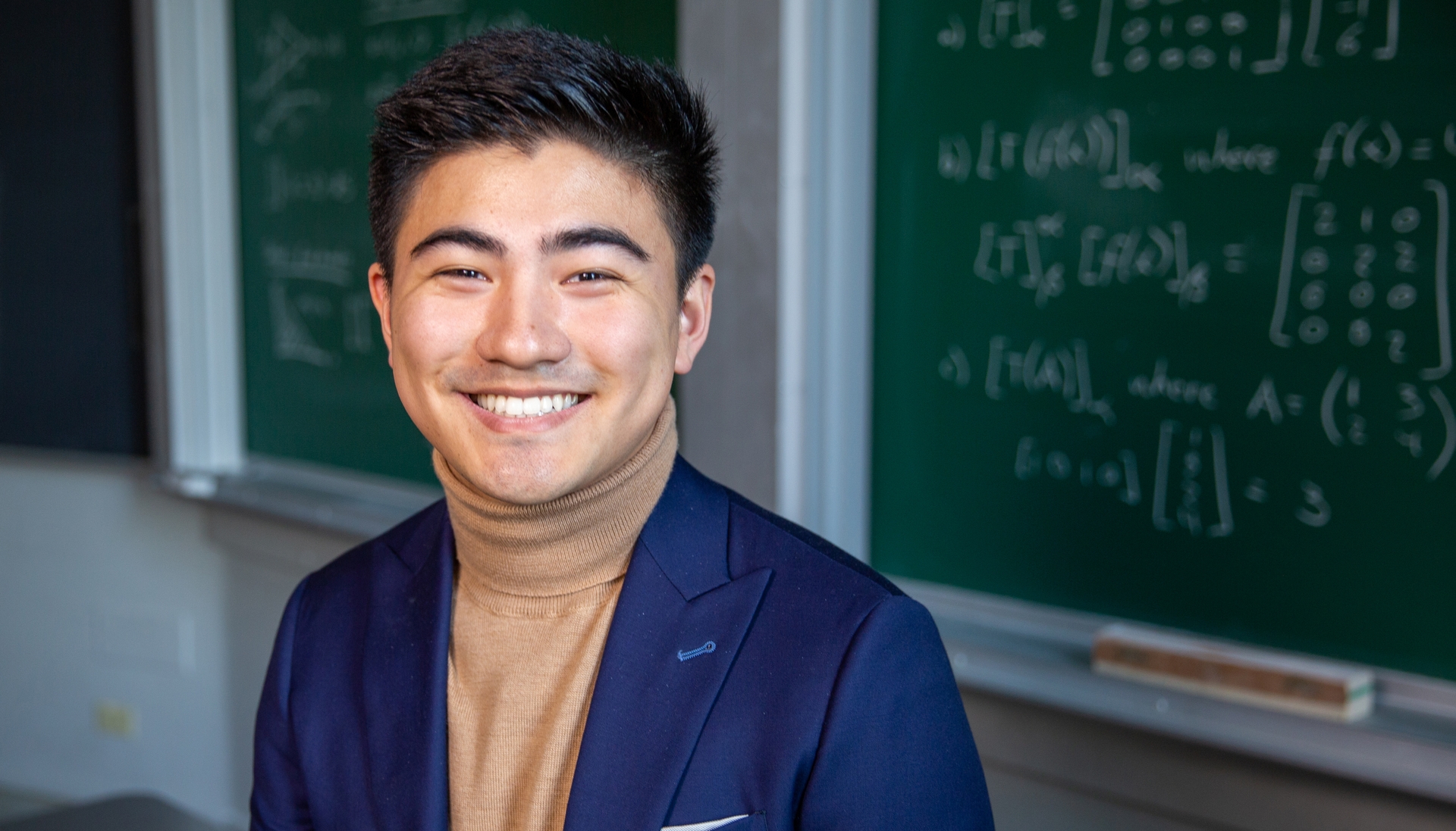 smiling young man in turtelneck and jacket standing in front of a chalkboard with math equations