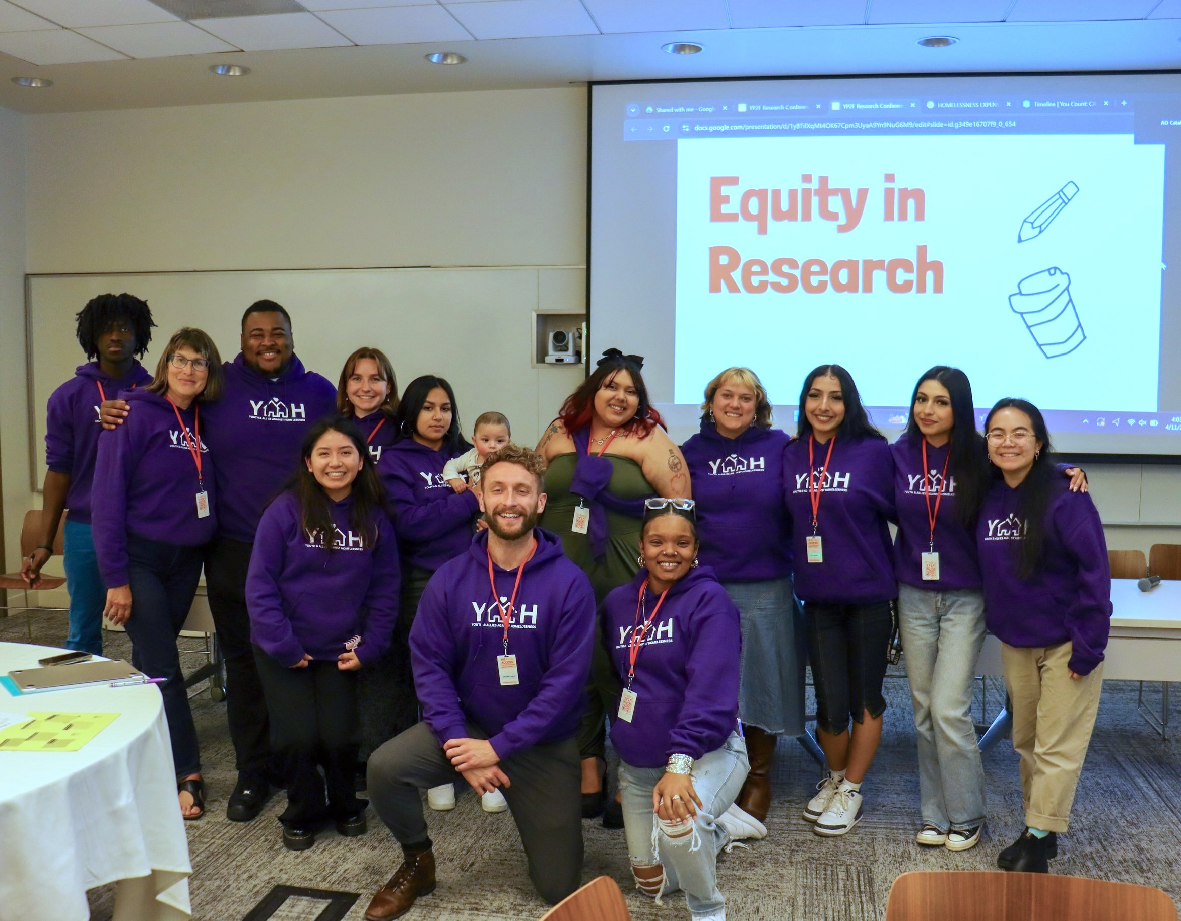 A group of 14 people who are involved in Coco Auerswald's project involving young people who experienced homelessness in the research process stand in front of a screen that reads 