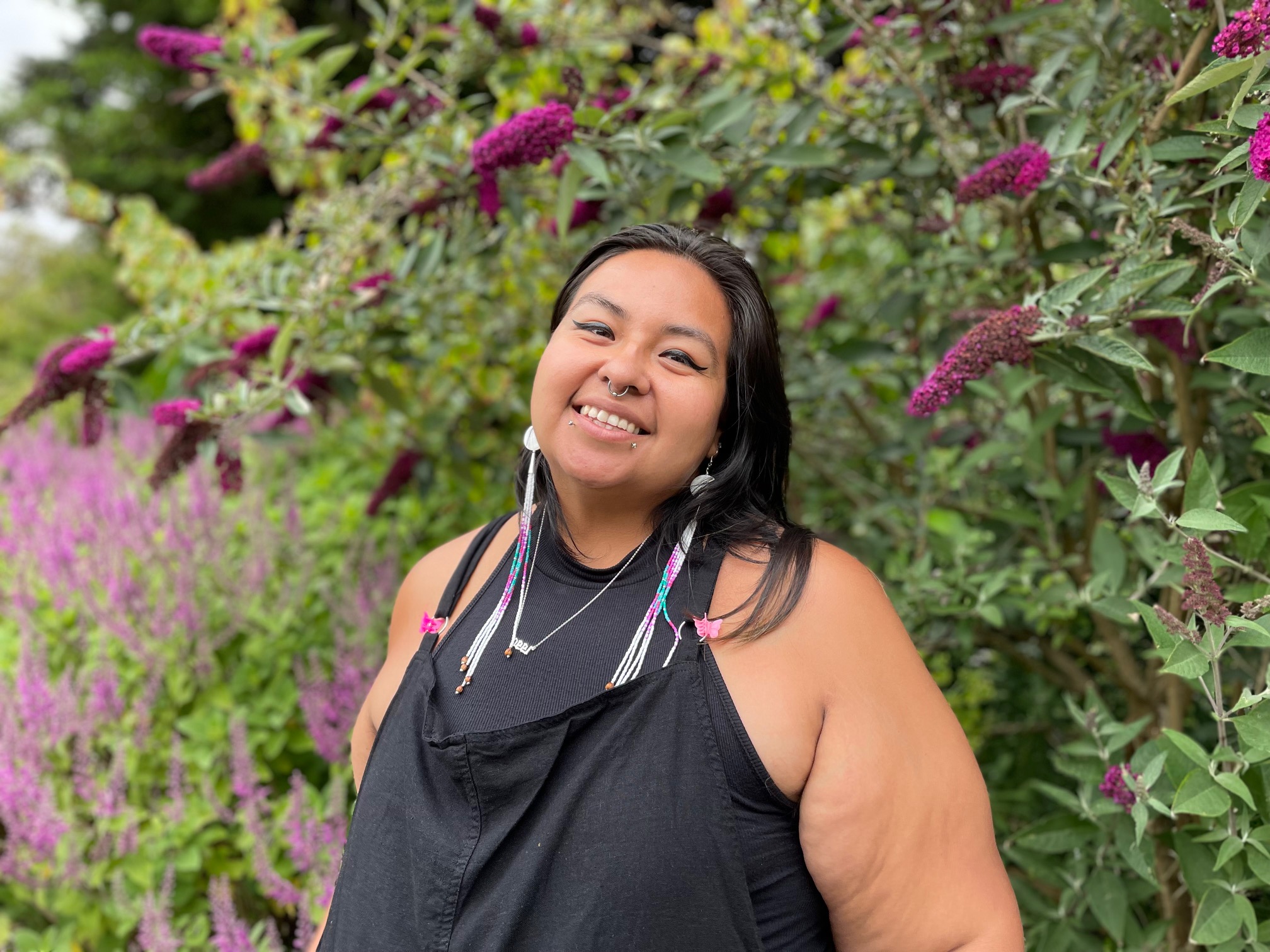 A portrait of Joanna Villegas, pictured before a background of green plants with purple flowers.