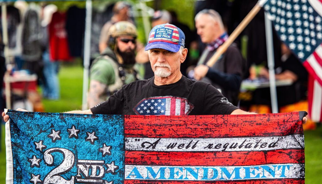 A bearded man in a baseball cap holds a U.S. flag that bears the words "A well regulated 2nd Amendment"