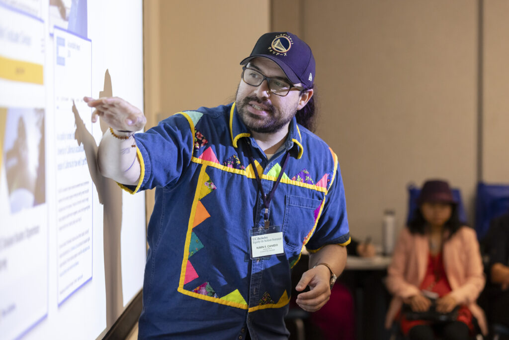 A man giving a presentation points to a screen on the wall where an image is projected that describes the subject he's talking about.