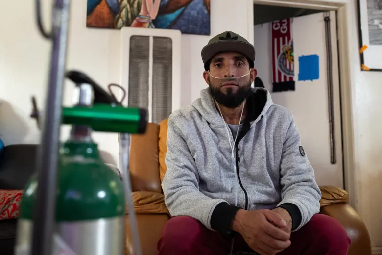 A bearded Latino man in a baseball cap looks solemnly at the camera. An oxygen cannula runs from a green tank into his nose.
