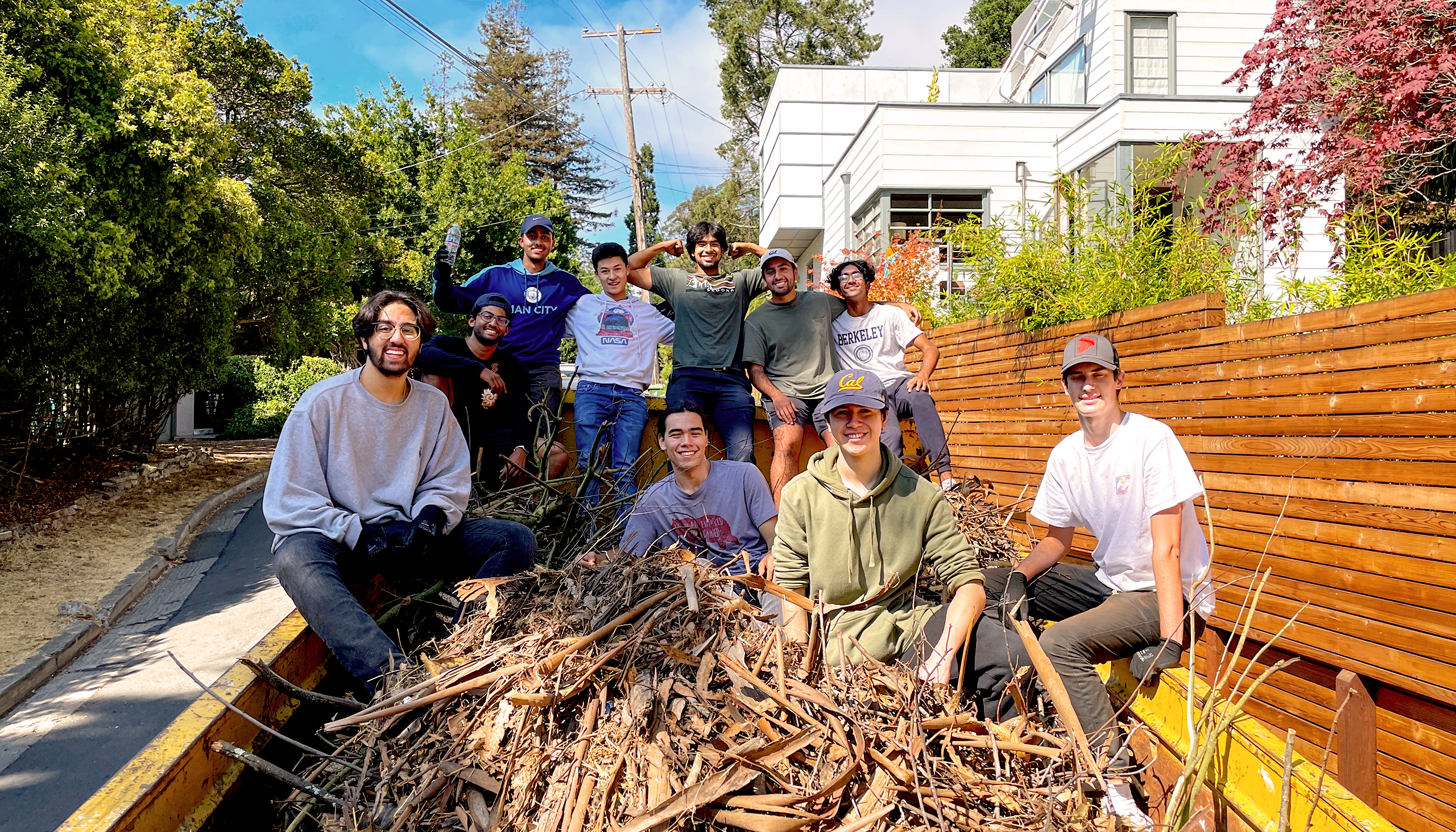 A group of students sits in a large yellow disposal bin that is filled with small twigs and branches. The bin is on a hilly street next to a white house.