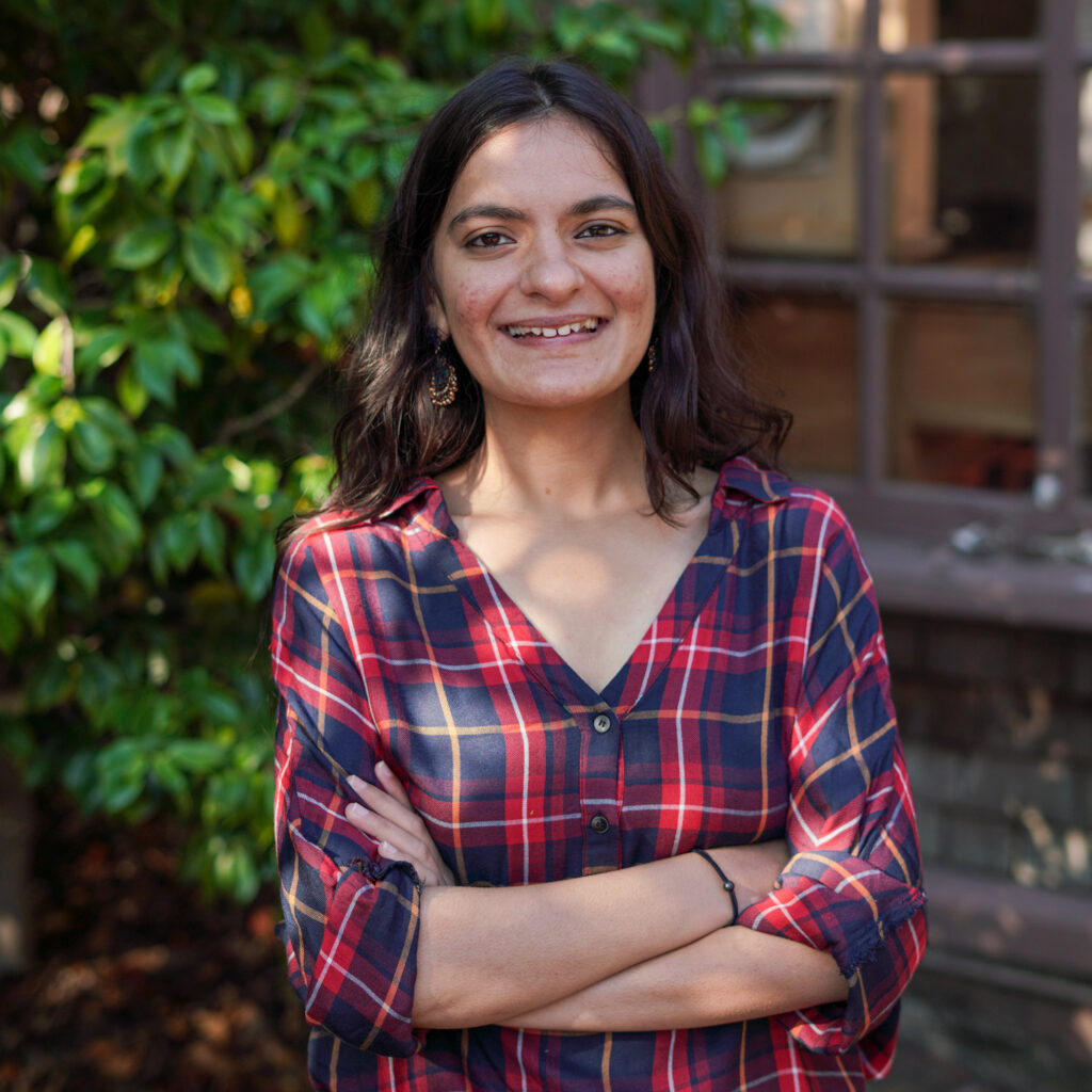 an Indian woman wearing a plaid shirt smiles at the camera in this headshot. She stands in front of foliage and has her arms crossed.