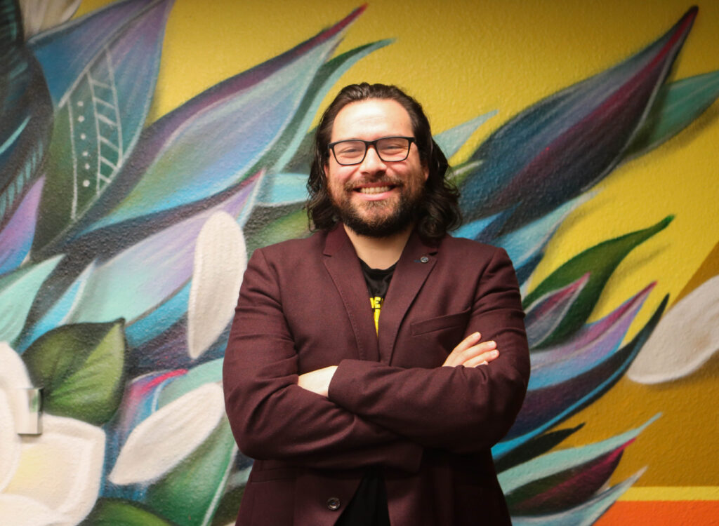 A man stands with his arms folded across his chest in front of a colorful mural of flowers with pointed leaves. He is smiling and wearing a rust-colored blazer, T-shirt, eyeglasses and has a beard and mustache.