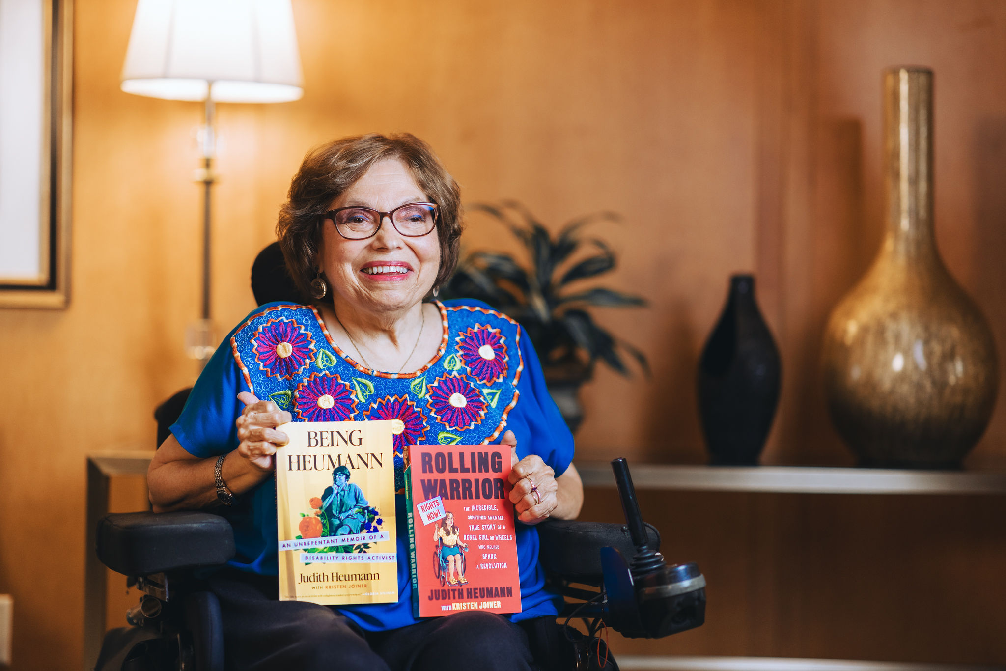 A photo of Judy Heumann holding her two books, Being Heumann and Rolling Warrior. Judy is a woman with short brown hair who uses a wheelchair. She is wearing glasses, a bright blue shirt with pink embroidered flowers, and black pants. The book covers are yellow and red.