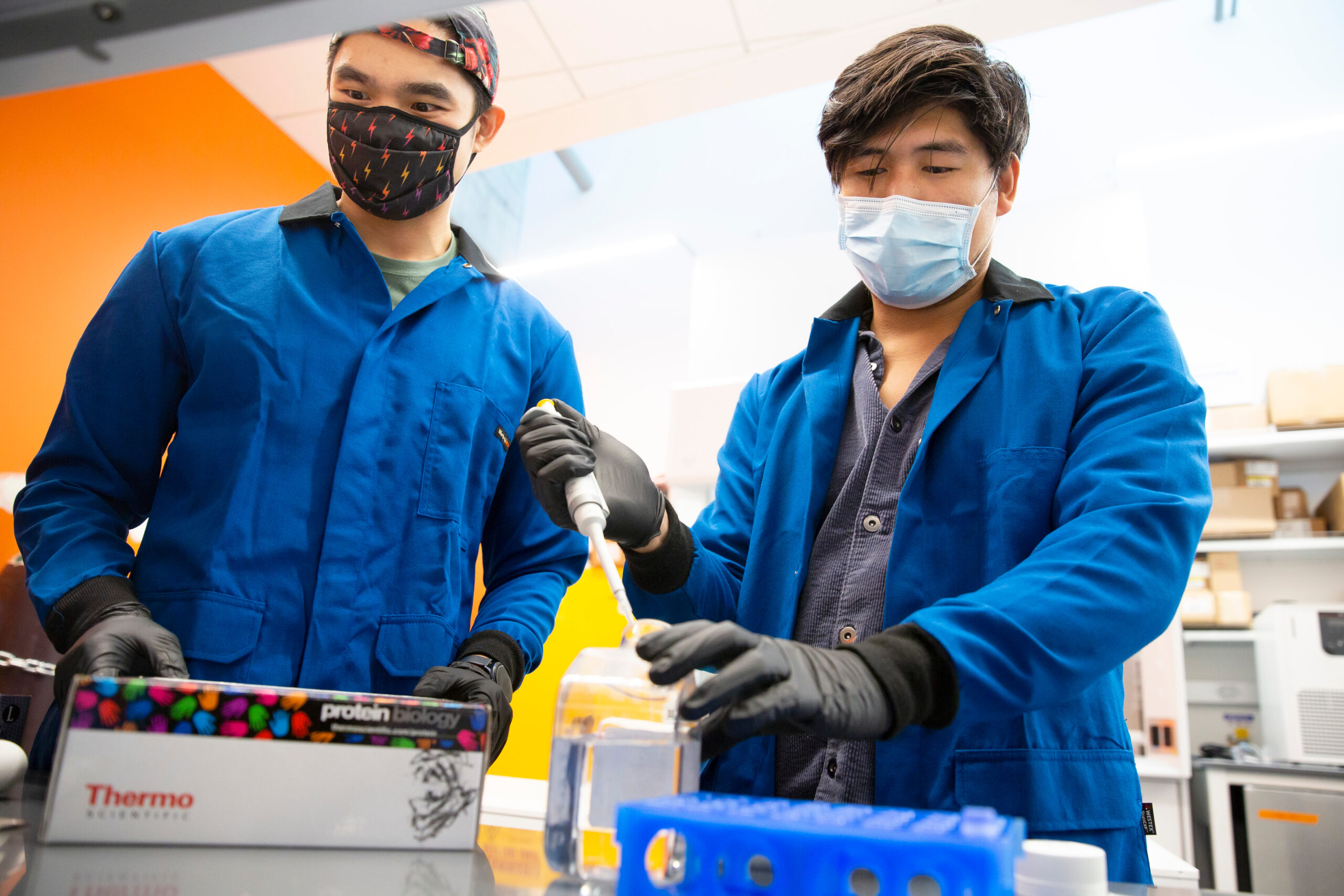 Two people wearing blue lab coats and masks sqeeze a tube at a lab bench into a vial.