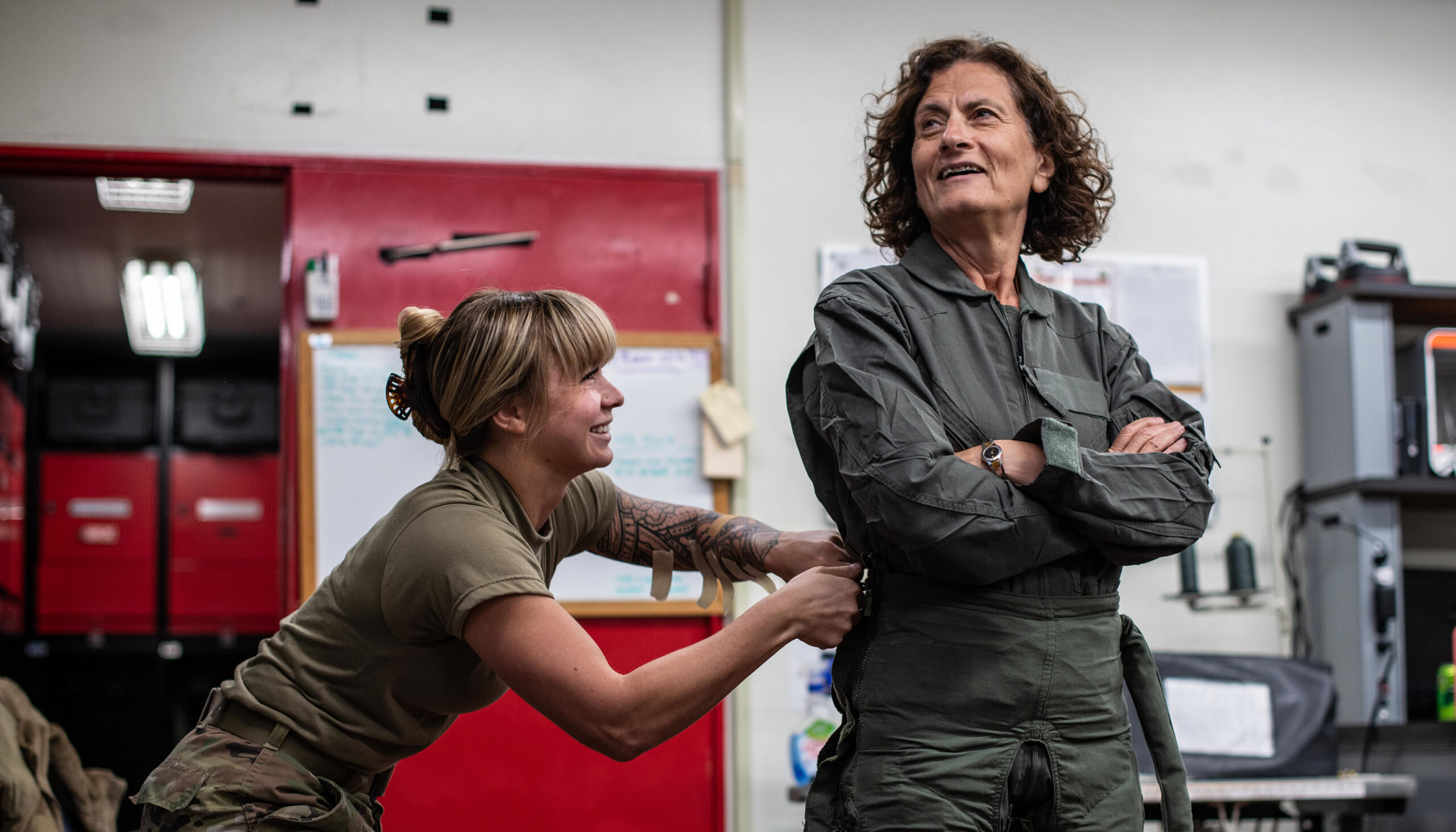 a young woman cinches up the green flight suit of an older woman